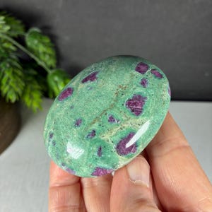 May include: A polished, oval-shaped zoisite stone featuring a vibrant green base color with embedded, deep magenta-colored ruby inclusions scattered across the smooth, glossy surface of the mineral specimen.