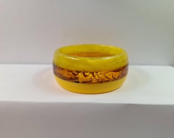 May include: A yellow resin bracelet with a band of amber-colored, textured material. The bracelet is a thick, rounded band, with a glossy finish. The amber band is encased between two layers of yellow resin.