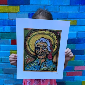 May include: A colorful painting of a woman with a halo above her head. The painting is framed and has the text "Love is the measure" written on it. The painting is being held by a person in front of a blue brick wall.