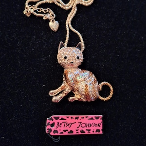 Può includere: Collana con pendente a forma di gatto dorato con un design di strass scintillanti. Il pendente a forma di gatto ha occhi neri e naso rosa. La collana include un piccolo ciondolo a cuore e un'etichetta con "BETSEY JOHNSON".