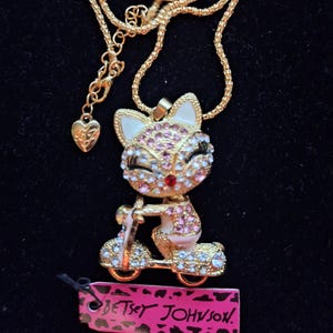Può includere: Una collana dorata con un ciondolo a forma di volpe su uno scooter, impreziosita da cristalli rosa e trasparenti. La volpe ha un naso di gemma rossa. La collana include un ciondolo a forma di cuore e un'etichetta del marchio "Betsey Johnson".