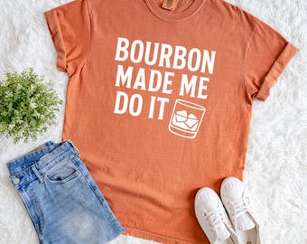 Peut inclure: Un t-shirt orange avec l'inscription blanche "BOURBON MADE ME DO IT" et un dessin de verre à whisky. Il est associé à un jean bleu et des baskets blanches, le tout sur un fond blanc duveteux.