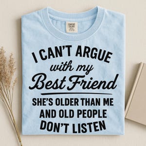 May include: Light blue Comfort Colors t-shirt with the text "I CAN'T ARGUE with my Best Friend SHE'S OLDER THAN ME AND OLD PEOPLE DON'T LISTEN" in black. The shirt is folded on a light beige surface.