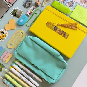 May include: A collection of stationery items, including a yellow notebook with a tassel bookmark, a teal pencil case, pens, highlighters, stickers, and a paperclip. The items are arranged on a light green surface.