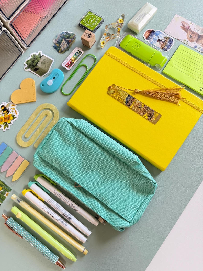 May include: A collection of stationery items, including a yellow notebook with a tassel bookmark, a teal pencil case, pens, highlighters, stickers, and a paperclip. The items are arranged on a light green surface.