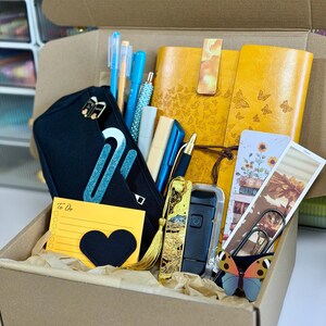 May include: A brown cardboard box filled with stationery items. Includes a black pencil case, yellow notebooks with butterfly designs, pens, a to-do list, and decorative paperclips. The box is lined with brown paper.