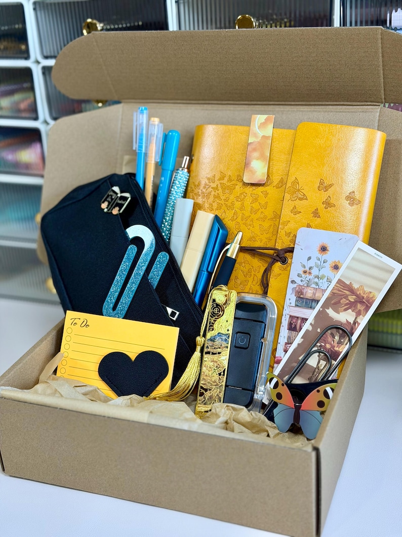 May include: A brown cardboard box filled with stationery items. Includes a black pencil case, yellow notebooks with butterfly designs, pens, a to-do list, and decorative paperclips. The box is lined with brown paper.