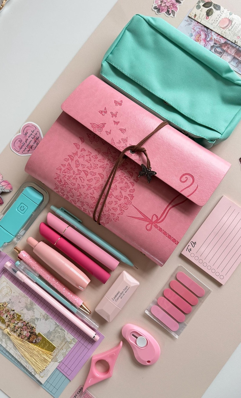 May include: A collection of pink and teal stationery items, including pens, a notebook with butterfly designs, a pencil case, sticky notes, and a pink utility knife. The items are arranged on a light beige surface.