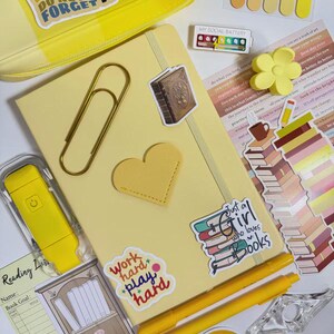 May include: A collection of stationery items in shades of yellow, including a notebook, pens, stickers, and a pencil case. The notebook has a gold heart and a large paper clip. Stickers include text like "Work hard play hard" and "Just a girl who loves books."