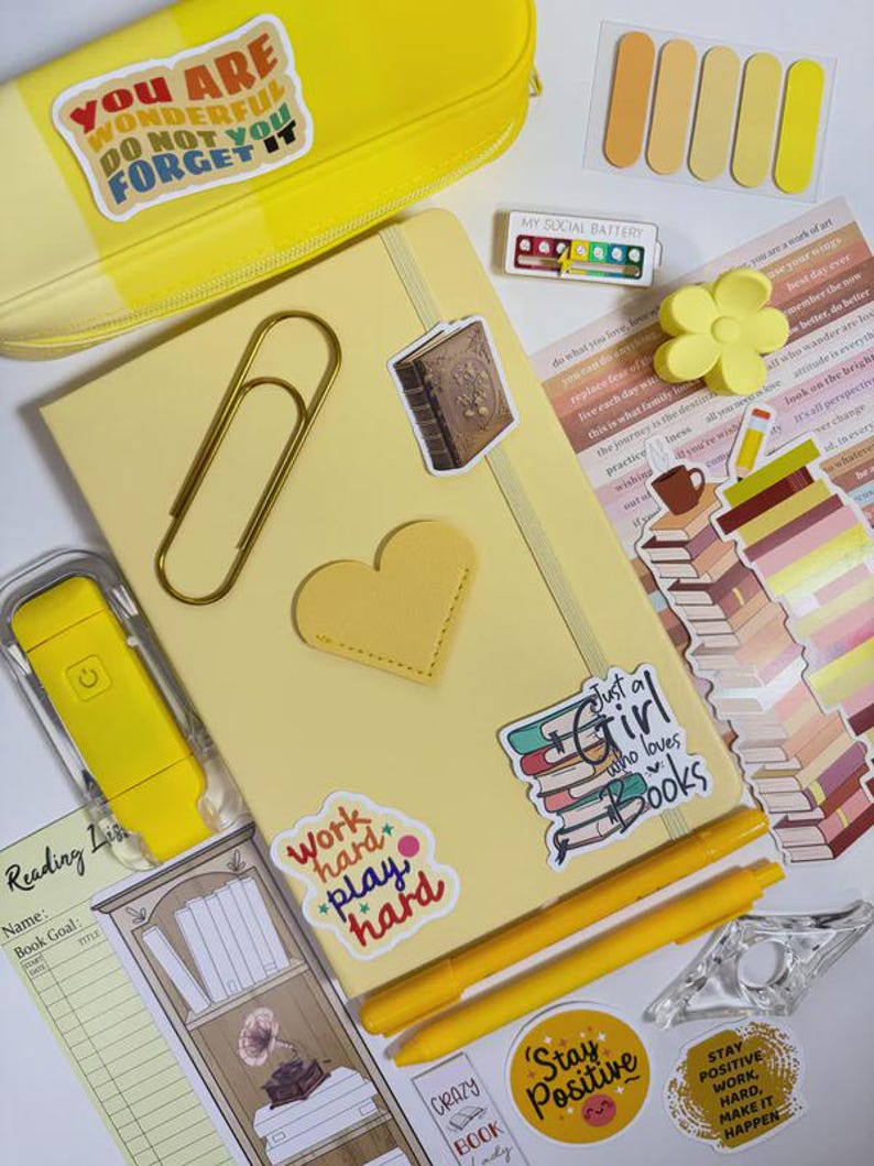 May include: A collection of stationery items in shades of yellow, including a notebook, pens, stickers, and a pencil case. The notebook has a gold heart and a large paper clip. Stickers include text like "Work hard play hard" and "Just a girl who loves books."