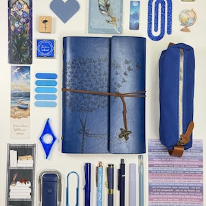 May include: A collection of blue stationery and accessories, including a journal, pens, a pencil case, bookmarks, and stickers. The journal is leather-bound with a butterfly design. The items are arranged on a white surface.