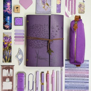 May include: A collection of purple stationery items, including a journal with butterfly designs, pens, a pencil case, stickers, and a bookmark. The items are arranged on a white surface, creating a visually appealing layout for journaling and creative projects.