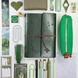 May include: An assortment of stationery and accessories in shades of green, including a journal, pencil case, pens, and a wallet. A metal bookmark with a feather design and the text "FANTASY FEATHERS AURORA" is also visible.