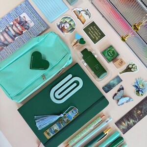 May include: An assortment of stationery items in shades of green and blue, including pens, a notebook, a pencil case, stickers, and a bookmark. The notebook has a large paperclip design. The image also includes a small notepad with "To Do" written on it.