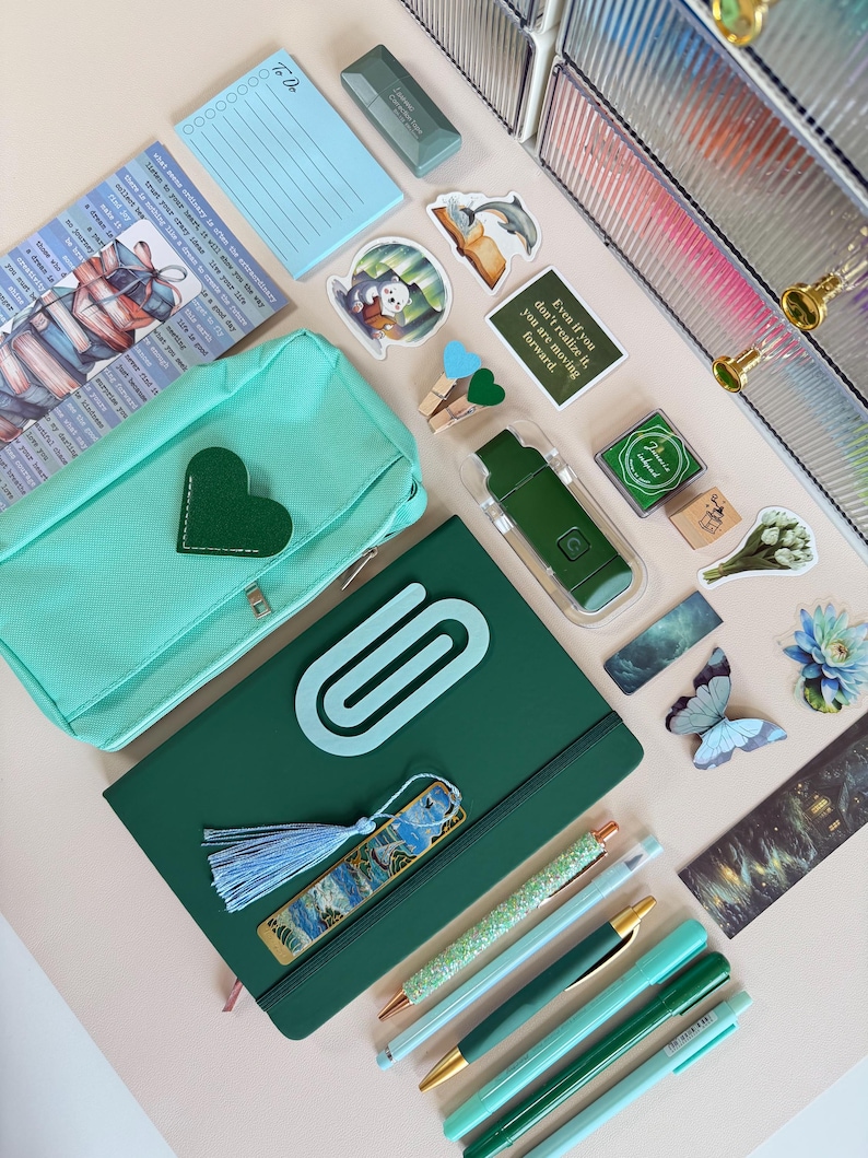 May include: An assortment of stationery items in shades of green and blue, including pens, a notebook, a pencil case, stickers, and a bookmark. The notebook has a large paperclip design. The image also includes a small notepad with "To Do" written on it.