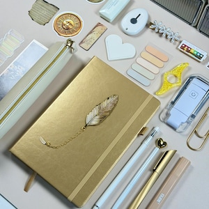 May include: A gold-colored journal with a feather charm, surrounded by stationery items. Includes pens, a pencil case, sticky notes, and decorative stickers. The items are arranged on a light pink surface.