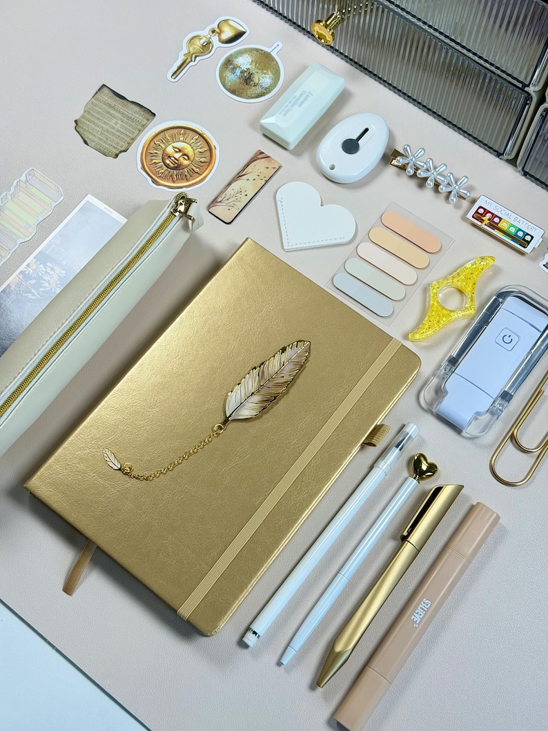 May include: A gold-colored journal with a feather charm, surrounded by stationery items. Includes pens, a pencil case, sticky notes, and decorative stickers. The items are arranged on a light pink surface.
