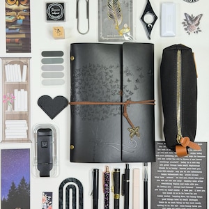 May include: An assortment of stationery items, including a black journal with butterfly details, pens, bookmarks, and stickers. A black pencil case and a feather-shaped metal bookmark are also visible. The items are arranged on a white surface.