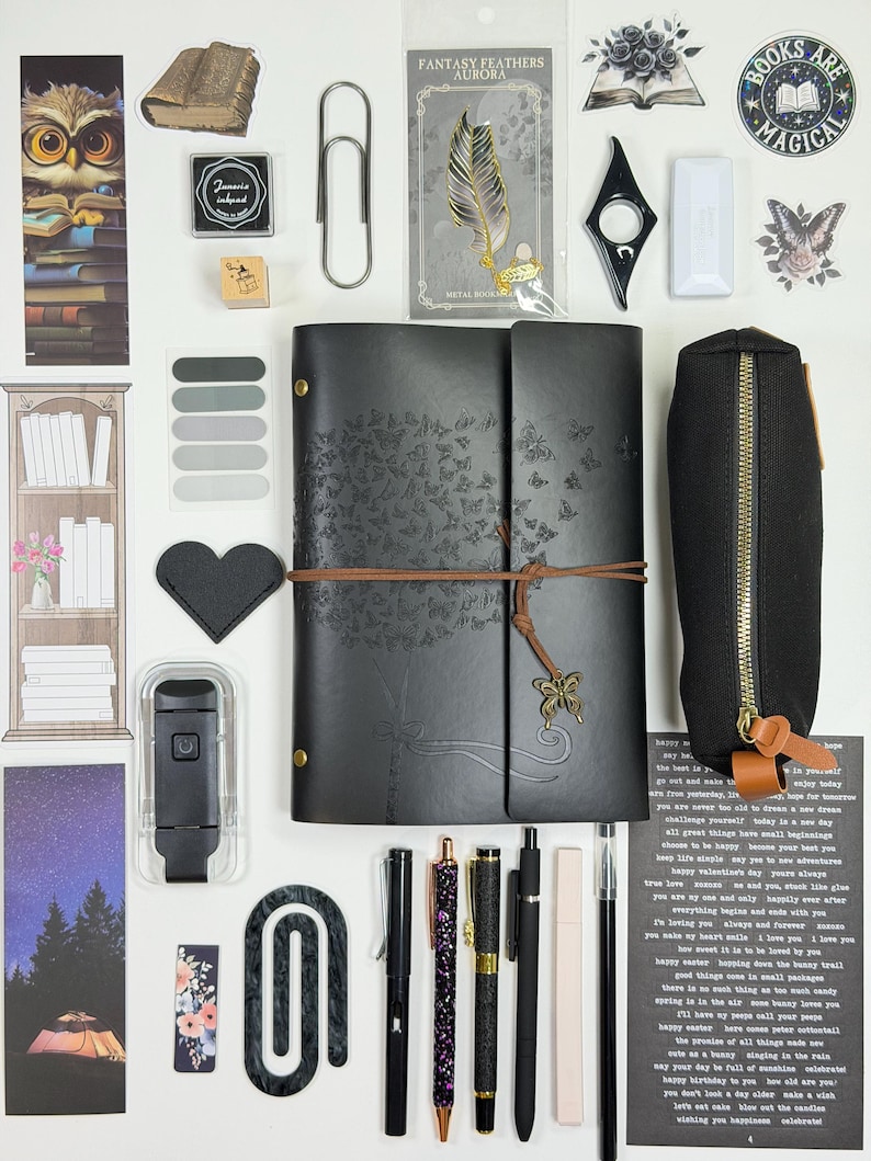 May include: An assortment of stationery items, including a black journal with butterfly details, pens, bookmarks, and stickers. A black pencil case and a feather-shaped metal bookmark are also visible. The items are arranged on a white surface.