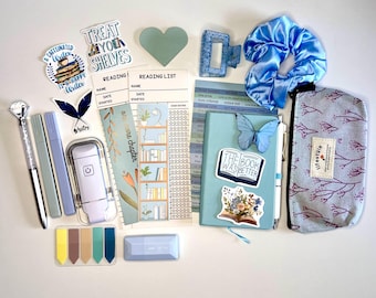 May include: A collection of stationery and accessories in shades of blue, lavender, and white. Includes a pen, highlighters, stickers, bookmarks, a scrunchie, a hair clip, a notebook, and a pencil case. The notebook has a butterfly and a sticker that says "THE BOOK WAS BETTER".