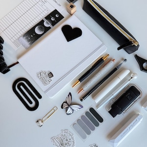 May include: An assortment of stationery items, including a white notebook with a black heart, pens, stickers, and a black pencil case with a gold zipper. The items are arranged on a white surface, with a reading list and bookmark visible.