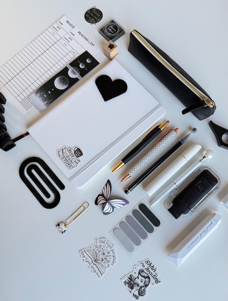 May include: An assortment of stationery items, including a white notebook with a black heart, pens, stickers, and a black pencil case with a gold zipper. The items are arranged on a white surface, with a reading list and bookmark visible.