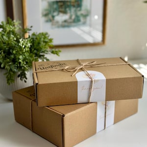 May include: Two stacked, rectangular brown cardboard boxes tied with twine. Each box has a white band with the words "thank you!" in gold script. The top box also has the word "Unifindit" printed on it.