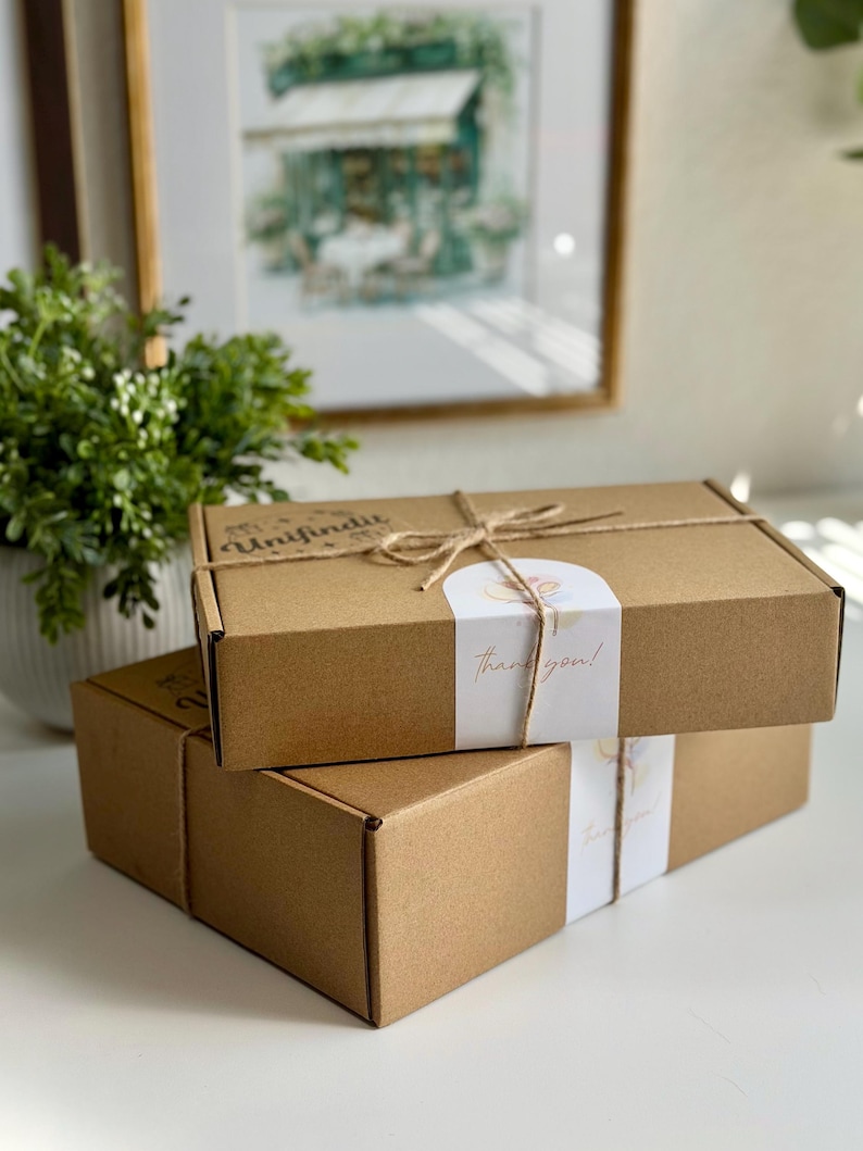 May include: Two stacked, rectangular brown cardboard boxes tied with twine. Each box has a white band with the words "thank you!" in gold script. The top box also has the word "Unifindit" printed on it.