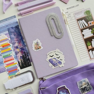 May include: A collection of purple and lavender stationery items, including pens, a notebook, stickers, and a pencil case. The notebook has a large silver paperclip. Stickers include the text "Books are Magic". A reading list and a book light are also present.