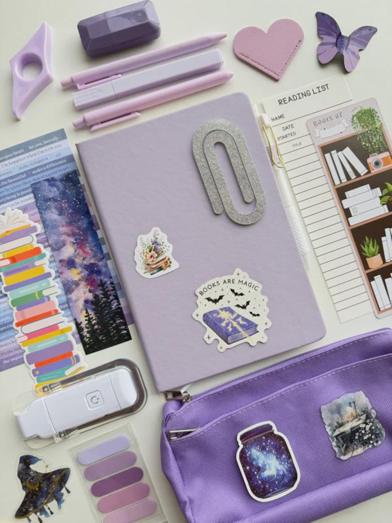 May include: A collection of purple and lavender stationery items, including pens, a notebook, stickers, and a pencil case. The notebook has a large silver paperclip. Stickers include the text "Books are Magic". A reading list and a book light are also present.