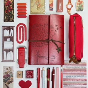 May include: A collection of stationery and decorative items, including a red journal with butterfly and tree designs, pens, stickers, and a pencil case. The items are arranged on a white surface, with a variety of colors and textures.