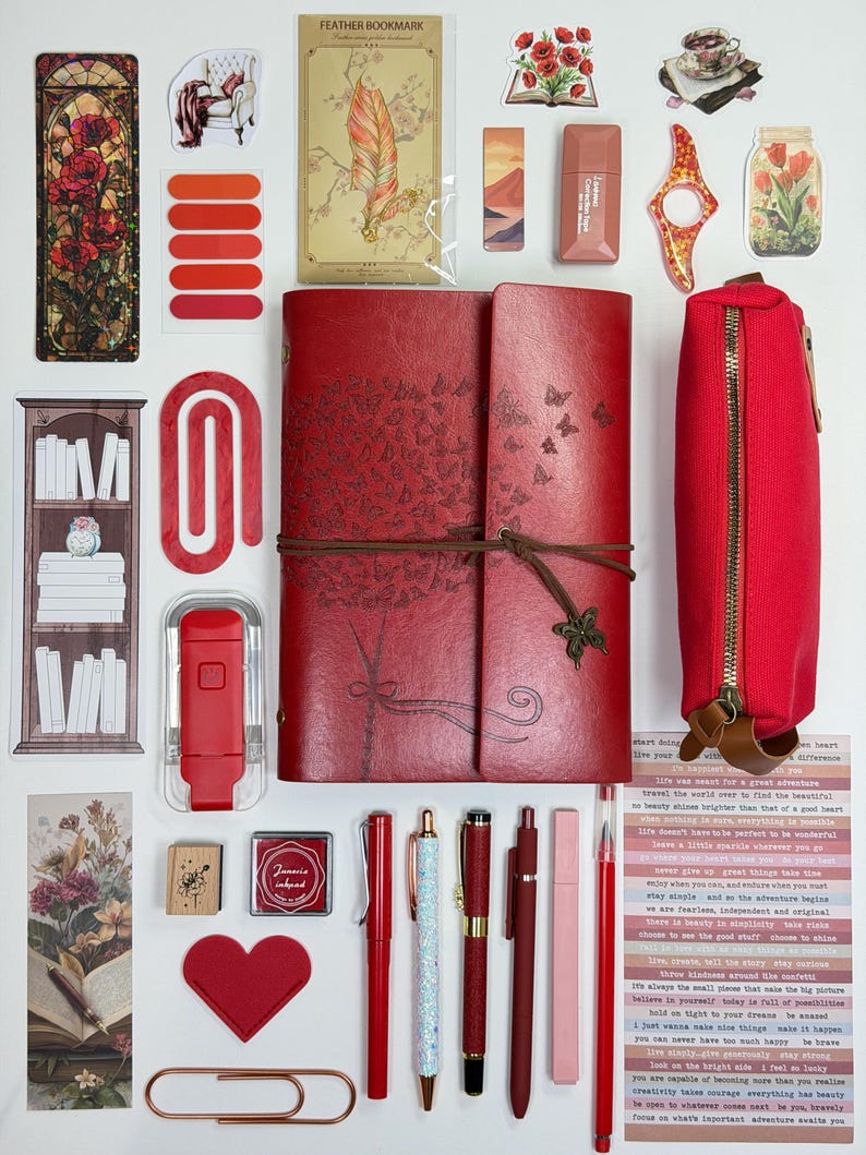 May include: A collection of stationery and decorative items, including a red journal with butterfly and tree designs, pens, stickers, and a pencil case. The items are arranged on a white surface, with a variety of colors and textures.