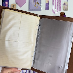 May include: Open brown leather binder with lined paper and a clear plastic sleeve. The binder is held open, revealing the interior. Decorative stickers and pens in shades of purple are visible in the background.