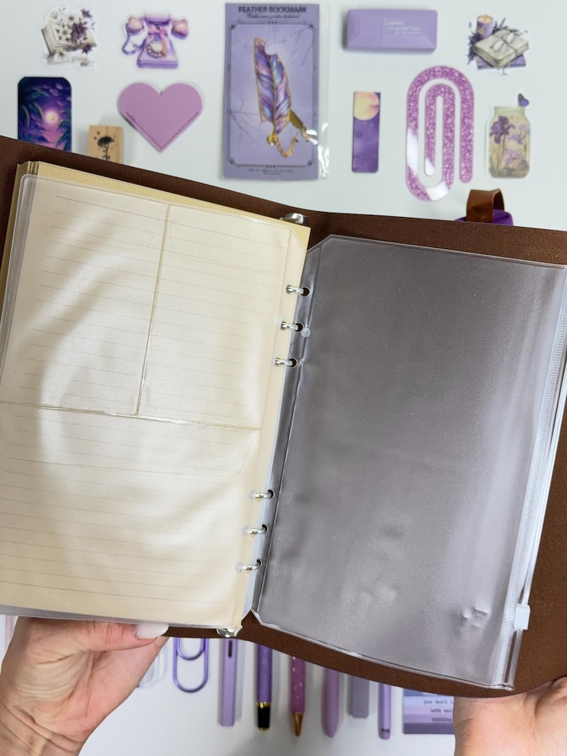 May include: Open brown leather binder with lined paper and a clear plastic sleeve. The binder is held open, revealing the interior. Decorative stickers and pens in shades of purple are visible in the background.
