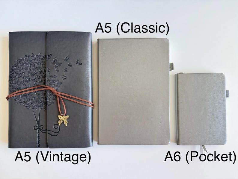 May include: Three notebooks of varying sizes are displayed. The largest, labeled "A5 (Vintage)", is dark gray with an embossed tree and butterfly design, secured with a brown cord and a butterfly charm. The "A5 (Classic)" and "A6 (Pocket)" notebooks are light gray.