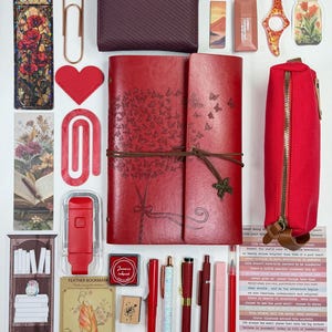 May include: A collection of red stationery and accessories, including a journal with butterfly designs, a wallet, pens, a pencil case, and stickers. Other items include a bookmark, a heart-shaped cutout, and a stamp. The items are arranged on a white surface.