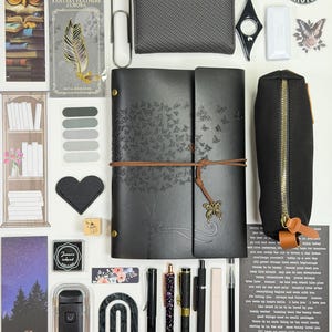 May include: An assortment of stationery items, including a black leather journal with butterfly and tree designs, pens, a pencil case, stickers, and a wallet. A metal bookmark with a feather design is also visible.