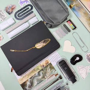 May include: An assortment of stationery items on a light green surface. Includes a black notebook with a gold feather design, pens, a pencil case, stickers, and a small black flashlight. The items are arranged for a creative workspace.