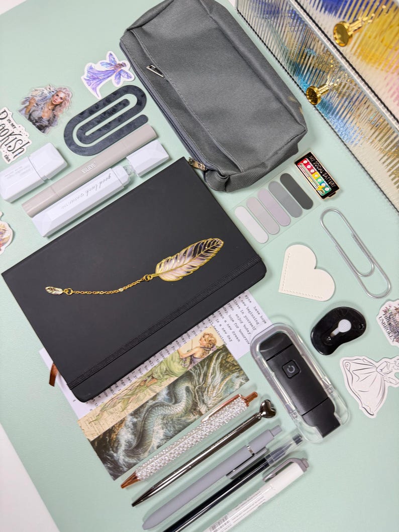 May include: An assortment of stationery items on a light green surface. Includes a black notebook with a gold feather design, pens, a pencil case, stickers, and a small black flashlight. The items are arranged for a creative workspace.