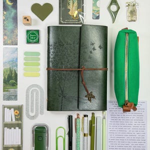 May include: An assortment of stationery and decorative items in shades of green and white. Includes a green journal, pens, stickers, a pencil case, and a metal bookmark with a feather design. The items are arranged on a white surface.