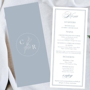 Puede incluir: Un menú de boda y una tarjeta a juego se muestran sobre una superficie blanca, con una ramita de flores blancas. El menú presenta un diseño formal con entrantes, platos principales y postres. La tarjeta tiene un diseño floral con las letras "C" y "R".