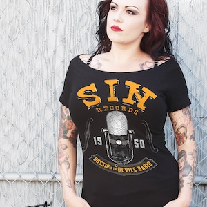 May include: A black off-the-shoulder t-shirt with a vintage-style graphic of a microphone and the text "SIN RECORDS 19 50 GOSSIP IS THE DEVILS RADIO".