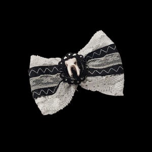 May include: A white lace bow with black ribbon accents and a decorative black center. The center features a small, realistic tooth in a black, ornate setting. The bow has a unique, gothic aesthetic.