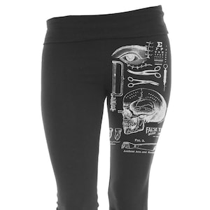 Puede incluir: Pantalones de yoga negros con una ilustración anatómica blanca de un ojo humano, un cráneo y herramientas quirúrgicas. El texto "FALSE TEETH BOUGHT" y "FIG. 3." también están incluidos en el diseño.