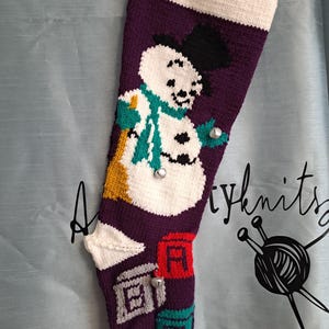 Può includere: Una calza natalizia lavorata a maglia di colore viola con un pupazzo di neve bianco che indossa un cilindro nero e una sciarpa verde acqua. Il design include blocchi alfabetici colorati in rosso, grigio e verde acqua con campanellini argentati.