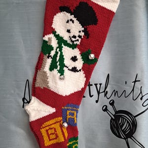 May include: A red knitted Christmas stocking featuring a white snowman wearing a black top hat and a green scarf. The stocking includes three small silver jingle bells attached to the front and a pattern of colorful alphabet blocks at the toe.