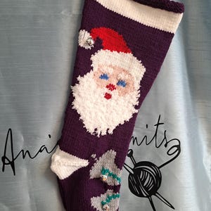 May include: A purple knitted Christmas stocking featuring a white and red Santa Claus face with blue eyes and a textured beard. The stocking has a white cuff and toe, with small silver bells attached to the Santa hat and two grey shapes below.