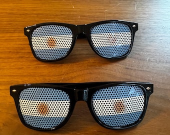 May include: Two pairs of black-framed sunglasses with the flag of Argentina displayed on the lenses. The flag features a light blue and white striped design with a golden sun in the center. The sunglasses are set against a wooden background.