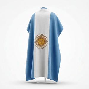 Puede incluir: Un poncho con la bandera argentina. El diseño incluye una franja blanca central con un emblema de sol dorado, flanqueada por paneles azul claro. La prenda se exhibe sobre un soporte blanco.
