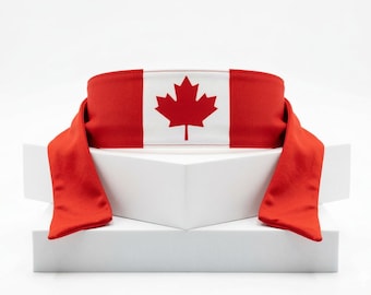 May include: A red and white headband featuring the Canadian flag. The headband has a red maple leaf on a white background, with red fabric extending from each side. The item is displayed on a white geometric structure.